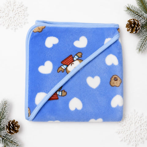 A folded blue plush blanket with a diagonal seam, decorated with white hearts, small brown bear faces, and a cute bear peeking out wearing a red striped hat. The blanket is styled in a flat-lay with pine branches and white snowflake decorations on a white background.