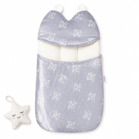 A baby footmuff in a light lavender-gray satin-like fabric with a white floral and star pattern, featuring a white inner lining and two white pom-poms. A small white plush star toy with embroidered facial features is placed to the left on a clean white background.