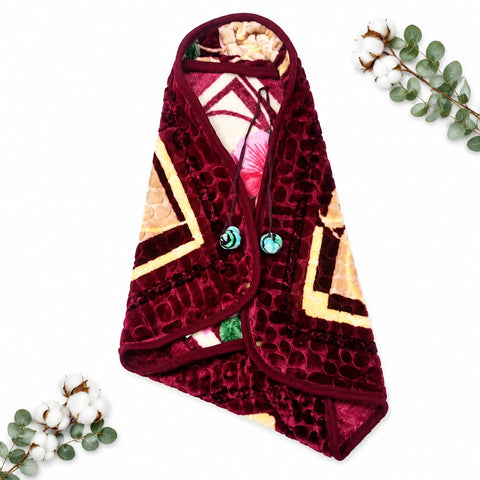 A plush fleece blanket with a geometric pattern, styled as a flat-lay on a white background with decorative cotton bolls and eucalyptus leaves. The blanket features a deep burgundy velvet exterior with a honeycomb texture, cream and yellow geometric panels, and turquoise buttons.