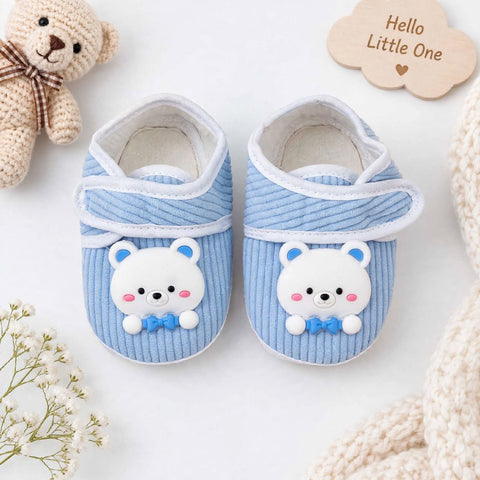 A pair of baby booties in light blue corduroy with white trim and a cute white bear face with a blue bow tie on the front. The booties are styled in a flat-lay with a plush teddy bear, a knit blanket, and a wooden cloud sticker that reads 'Hello Little One'.