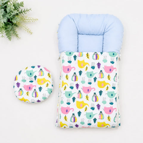 A baby footmuff and matching circular cushion set with a colorful pattern of pink teapots, yellow elephants, and green leaves on a white background, styled on a white surface with a green plant in the corner.
