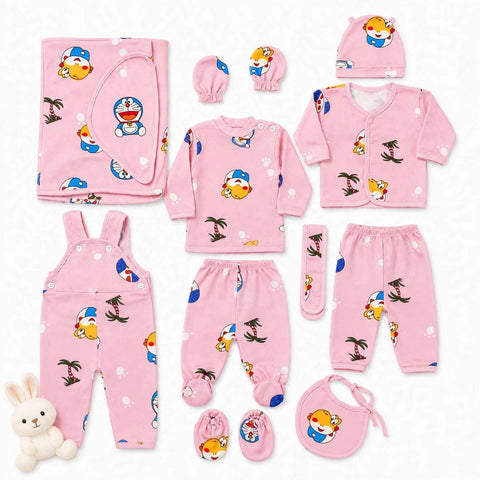 A pink baby clothing set arranged on a white background, featuring a long-sleeve top, cardigan, pants, overalls, footed socks, bib, hat, and booties, all printed with cartoon characters including Doraemon, a turtle, palm trees, and a small plush bunny.