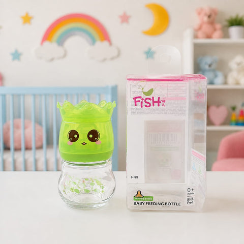 A green cartoon baby feeding bottle with a crown-shaped lid and a clear glass base, shown next to its retail packaging box on a white tabletop. The background is a softly blurred nursery setting with a crib, rainbow wall decor, and shelves.