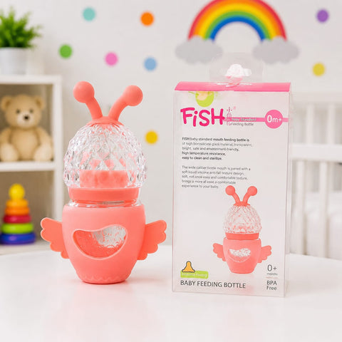 A pink baby feeding bottle with a clear textured top and wing-shaped base stands next to its retail box on a white surface. The box is labeled 'FISH' and 'Feeding Bottle' with age marking '0+'.