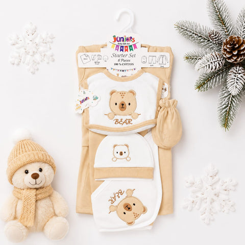 A baby starter set laid flat on a white background with holiday styling props. The set includes a cream and white bib with a teddy bear graphic, matching cream and white footmuff, cream and white booties, and a cream and white hat, all from Junior's Party.