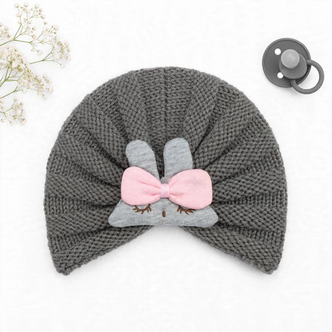 A gray knit baby hat with a bunny face applique and pink bow, shown on a white background with a pacifier and baby's breath flowers as props.