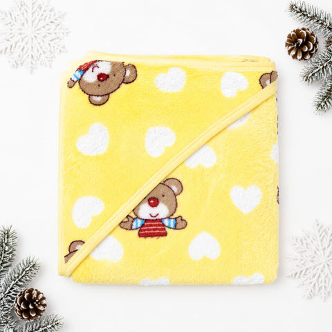 A yellow plush hooded towel blanket with a diagonal strap, decorated with white hearts and cartoon teddy bears wearing red and blue striped sweaters. The towel is styled in a flat-lay with pine branches and white snowflake decorations on a white background.