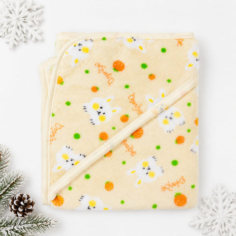 A folded cream-colored hooded baby blanket with a daisy and orange pattern, decorated with 'Daisy' text and small green dots, styled with pine branches, a pinecone, and white snowflakes on a white background.
