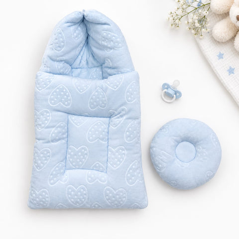 A light blue baby swaddle blanket with a hood and quilted heart pattern, shown flat-laid next to a matching round cushion and a small pacifier on a white background.