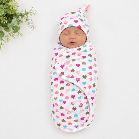 A sleeping newborn baby swaddled in a white muslin swaddle blanket with a colorful heart pattern, wearing a matching beanie hat, laid flat on a white background with a small green plant in the top left corner.