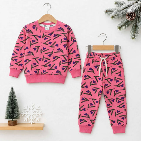 A children's two-piece outfit set consisting of a pink long-sleeve sweatshirt and matching jogger pants, both featuring a dark green leaf pattern with purple heart accents. The sweatshirt is shown on a wooden hanger on the left, and the pants on a metal hanger on the right, set against a clean white background with decorative holiday props.