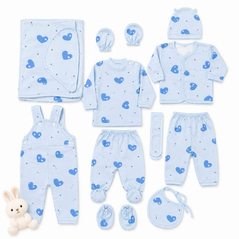 A baby clothing set laid out on a white background, including a light blue long-sleeve top, matching footed pants, overalls, a cardigan, a bonnet, a bib, a hat, a muslin cloth, and booties, all printed with a blue heart pattern. A small plush bunny toy is placed in the lower left corner.