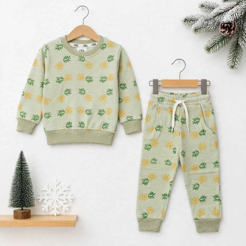 A children's two-piece outfit set consisting of a long-sleeve sweatshirt and jogger pants, both in a light sage green color with a yellow pineapple print. The sweatshirt is shown on a wooden hanger, and the matching pants are shown on a metal hanger, set against a white background with decorative holiday props.