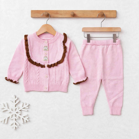 A toddler girl's two-piece knit outfit set consisting of a pink cable-knit cardigan with brown ruffle trim and matching pink knit pants, displayed on wooden hangers against a light textured background.