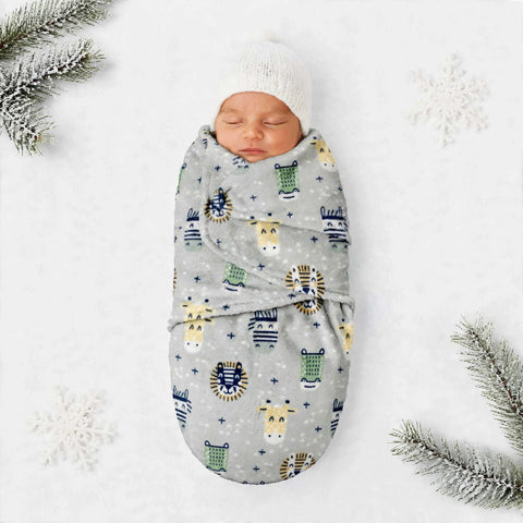 A sleeping newborn baby wrapped in a light gray blanket with a colorful animal print, wearing a white knit beanie, laid on a white background with decorative snowflakes and pine branches.