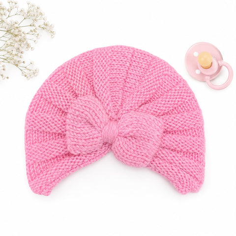 A pink knitted baby turban hat with a large matching bow, displayed on a white background with a pacifier and small white flowers as props.