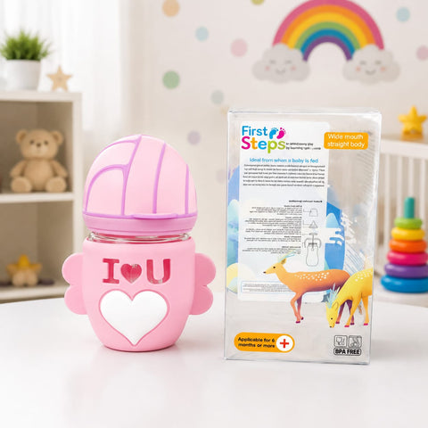 A pink baby bottle with a matching pink cap and a white heart on the front, shown next to its retail packaging box. The background is a softly blurred nursery setting with a rainbow, clouds, and a teddy bear.