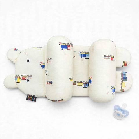 A baby support pillow designed in the shape of a bear, featuring a cream-colored fabric with a colorful cartoon train pattern. A small black fabric tag is attached near the bear's nose, and a blue pacifier is placed to the right on a clean white background.