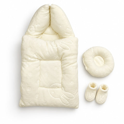 An off-white baby sleep sack with a hood and bunny motif, shown alongside a matching circular cushion and two booties on a clean white background.