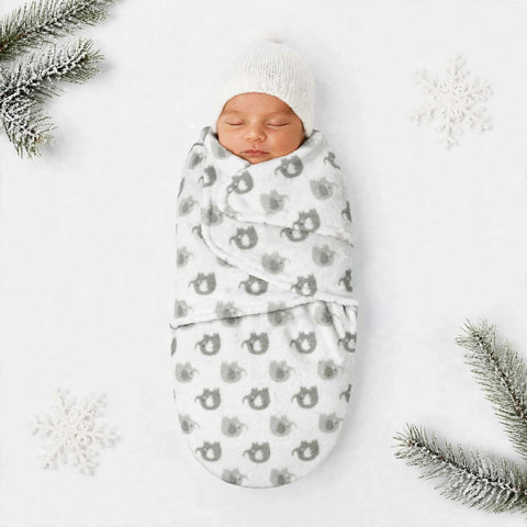 A sleeping newborn baby swaddled in a white blanket with a gray cat pattern, wearing a white knit hat, laid flat on a white background with decorative snowflakes and pine branches.