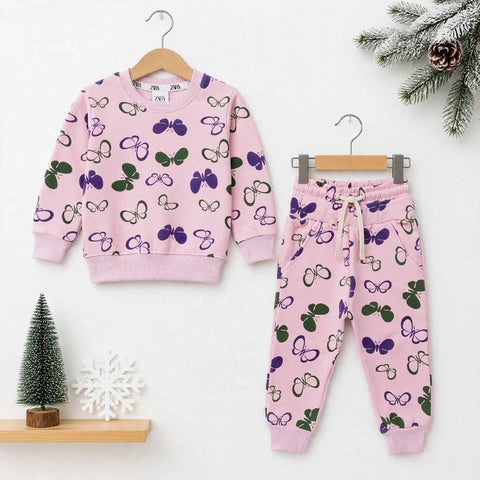 A children's two-piece lounge set consisting of a pink sweatshirt and matching jogger pants, both decorated with a purple and green butterfly pattern. The sweatshirt is shown on a wooden hanger on the left, and the pants on a metal hanger on the right, set against a clean white background with decorative holiday props.