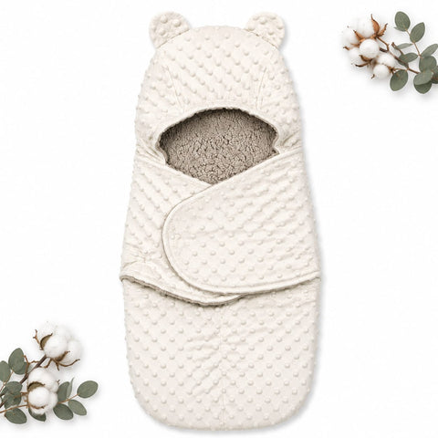 An off-white textured baby swaddle wrap with a hood featuring bear ears and a soft gray fleece lining, displayed on a white background with decorative cotton and eucalyptus sprigs.