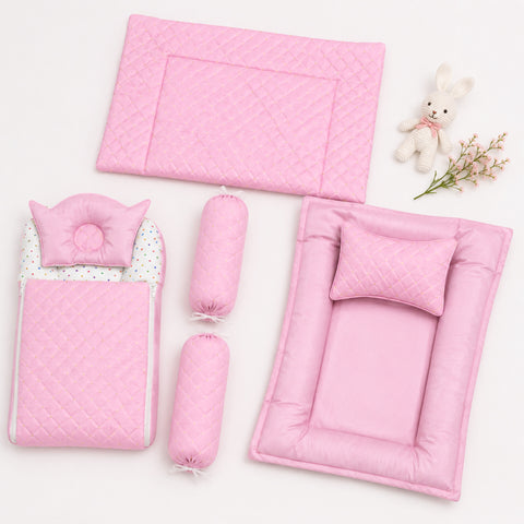 Pink quilted baby bedding set arranged on a white background, including a changing pad, mattress pad, two cylindrical bolster pillows, a small cushion, and a plush bunny toy with dried flowers.