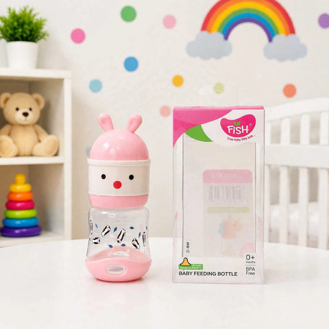 A pink and white baby feeding bottle with a bunny design next to its retail packaging box, set on a white table in a decorated nursery room.