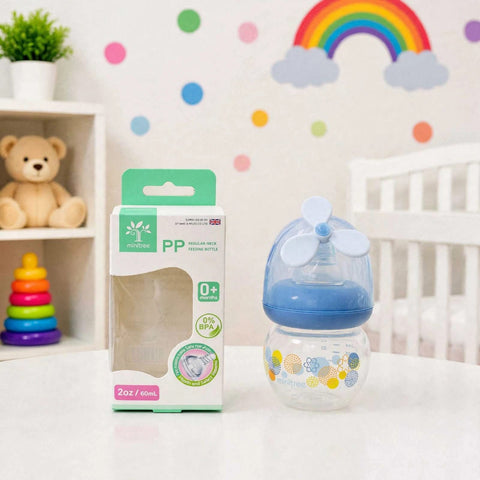 A baby bottle with a blue and white propeller spout and yellow patterned base, shown next to its green and white retail box on a white tabletop in a nursery setting.