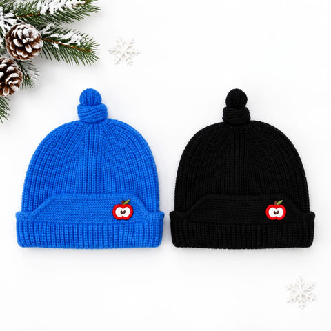 Two ribbed knit pom-pom beanies on a white background, one bright blue and one black, each with a small red apple patch on the front. Decorative pine branches, snow, and snowflakes appear around the hats.