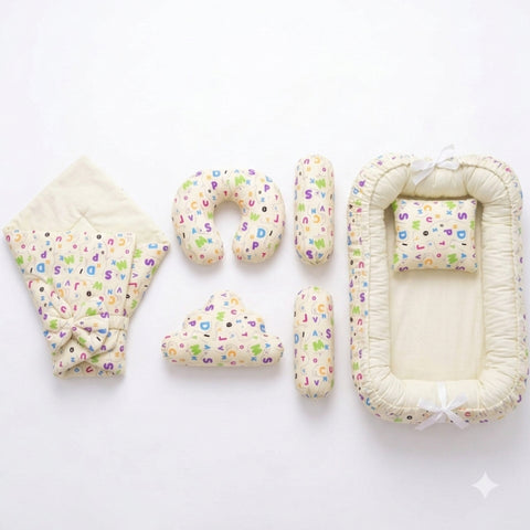 A baby bedding set laid out on a white background, including a quilted blanket with a colorful alphabet print and bow, a U-shaped pillow, two cylindrical pillows, a cloud-shaped pillow, and a bassinet cover with matching printed fabric and white ribbon ties.