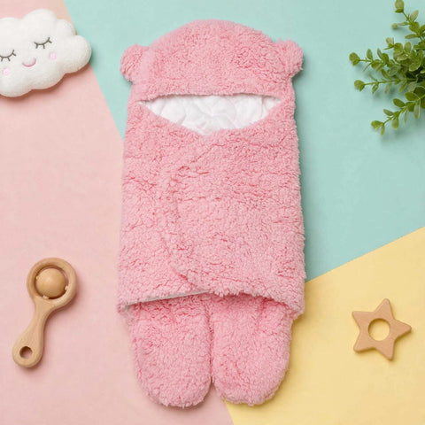 A pink plush baby foot warmer or bootie set laid flat on a pastel geometric background with a cloud toy, wooden rattle, greenery, and a star teether around it.