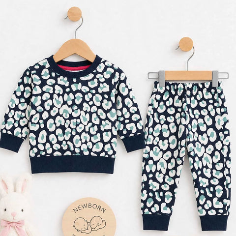 A baby clothing set displayed on wooden hangers against a white background, featuring a long-sleeve top and matching pants in navy blue with a white and mint green abstract pattern. A plush white bunny toy, a round wooden sign reading 'Newborn Essentials,' and a pair of white booties are arranged below.