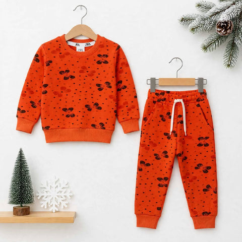 A children's two-piece outfit set consisting of an orange sweatshirt and matching jogger pants, both printed with small black motifs and dotted patterns. The sweatshirt is shown on a wooden hanger on the left, and the pants on a gray hanger on the right, styled against a white background with holiday props including a miniature tree, a snowflake, and pine branches.