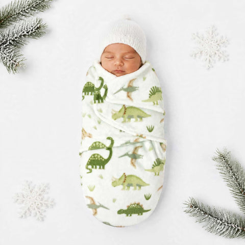 A sleeping newborn baby swaddled in a white fleece wrap with a green dinosaur pattern, wearing a white knit beanie, styled on a white background with decorative snowflakes and pine branches.