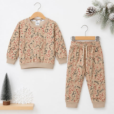 A children's two-piece pajama set hanging on wooden hangers against a white background, featuring a beige sweatshirt and matching joggers with an all-over abstract print in black, red, and orange. Decorative holiday props including a small tree, snowflake, pinecones, and evergreen branches are arranged around the set.