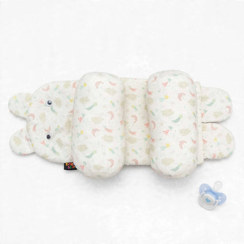 A plush baby support pillow shaped like a bunny, featuring a soft cream-colored fabric with a pastel rainbow pattern of rainbows and clouds. A small black fabric tag is visible near the bunny's nose, and a blue pacifier lies to the right on a clean white background.