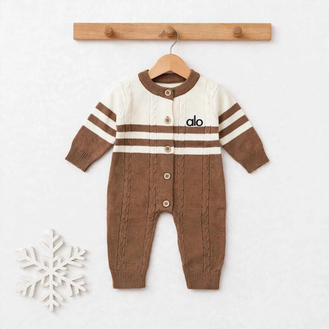 A baby knit cardigan jumpsuit in brown and cream, hanging on a wooden hanger from a wooden peg rack. The jumpsuit features a cream upper chest and shoulders with brown stripes, a brown button-front closure, and a brown cable-knit lower body.
