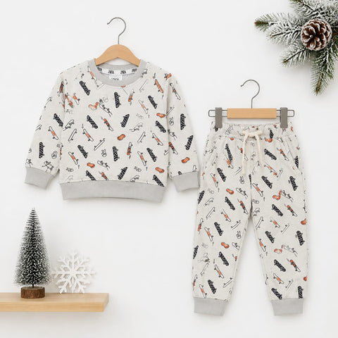 A children's two-piece clothing set featuring a long-sleeve sweatshirt and matching jogger pants, both in a warm off-white color with a playful print of black and orange winter-themed illustrations. The set is displayed on wooden hangers against a clean white background, decorated with a small artificial Christmas tree, a snowflake ornament, and a pine branch with a pinecone.