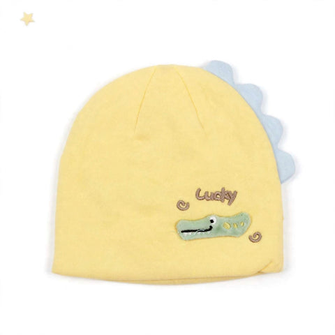 A pale yellow knit baby hat featuring a light blue scalloped dinosaur tail and an embroidered green crocodile face with the word 'Lucky' in brown script.