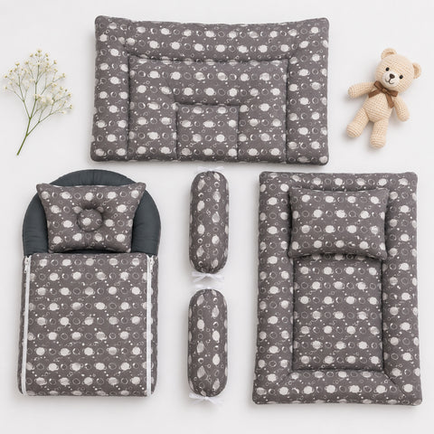 A baby bedding set arranged on a white background, including a changing pad, mattress, footmuff, two cylindrical bolsters, and a matching pillow, all in a gray fabric with a white cloud pattern. A small teddy bear and baby's breath sprig are placed as styling props.