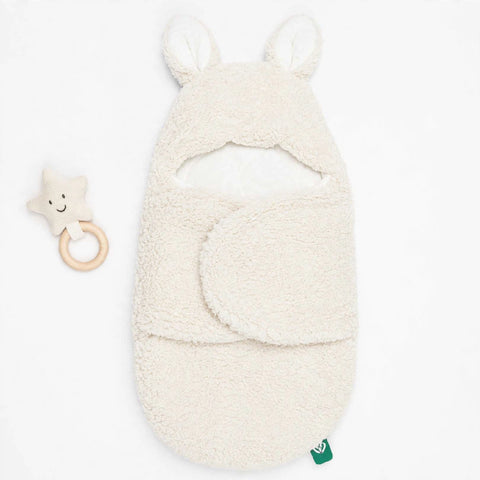 A cream-colored plush baby swaddle blanket with bunny ears on the hood, shown on a white background next to a matching star-shaped wooden teething ring.