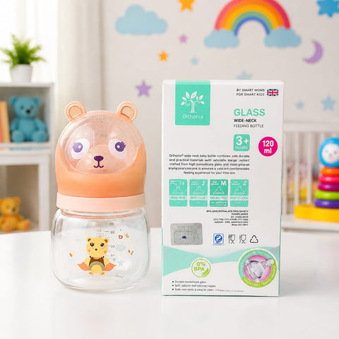 A baby feeding bottle with a bear face lid and a clear glass base, shown next to its product packaging box on a white surface against a decorated pastel background.