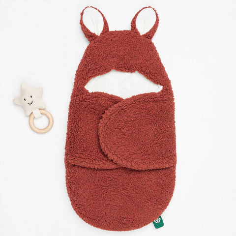 A terracotta-colored plush baby wrap or swaddle with a hood featuring pointed ears, shown on a white background next to a small cream star rattle.