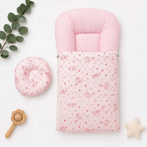 A pink baby footmuff with a printed pattern of cherubs and stars, shown alongside a matching pink circular cushion on a white background with decorative eucalyptus leaves, a wooden rattle, and a star toy.