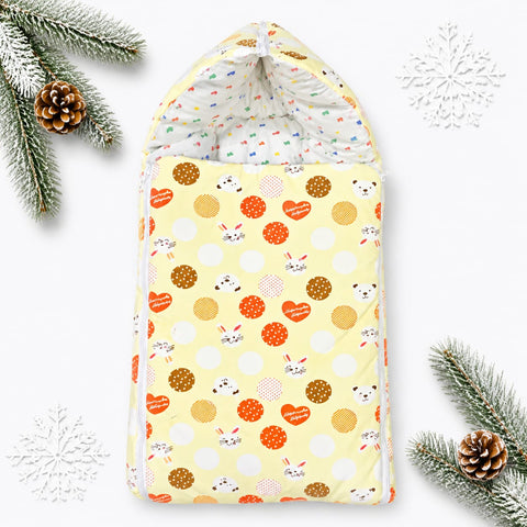 A baby swaddle blanket with a festive holiday print, featuring a pale yellow background with orange hearts, brown polka dots, white polka dots, and cute bunny faces. The swaddle is styled flat-lay with pine branches, pinecones, and white snowflake decorations on a clean white background.