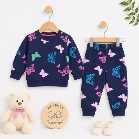 A baby clothing set displayed on wooden hangers against a white background, including a navy long-sleeve top and matching jogger pants with pink and blue butterfly prints. A cream teddy bear, a round wooden sign reading 'Newborn Essentials,' cream booties, and a small green leafy sprig are arranged around the set.
