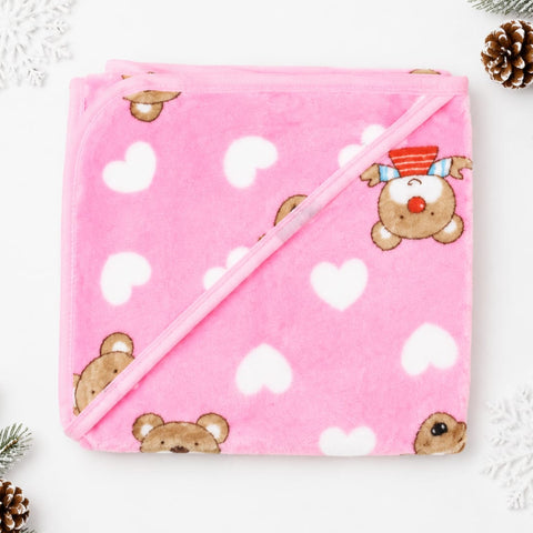 A folded pink hooded blanket with a white heart pattern and cute teddy bear faces, decorated with a small red and blue striped hat on one bear. The blanket is styled on a white background with decorative pinecones and snowflakes.