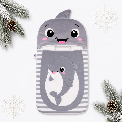 A plush baby sleeping bag or pouch with a shark hood and a cartoon shark graphic on the front, styled with pine branches and snowflakes on a white background.