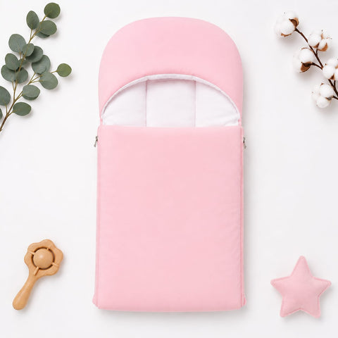 A light pink baby footmuff or swaddle bag with a white inner lining, styled in a flat-lay arrangement with a wooden rattle, green eucalyptus leaves, cotton bolls, and a pink star toy on a white background.